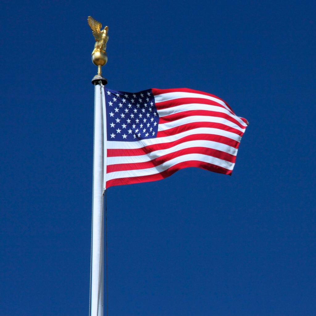 United States flag waving against a clear blue sky, topped with a golden eagle finial.